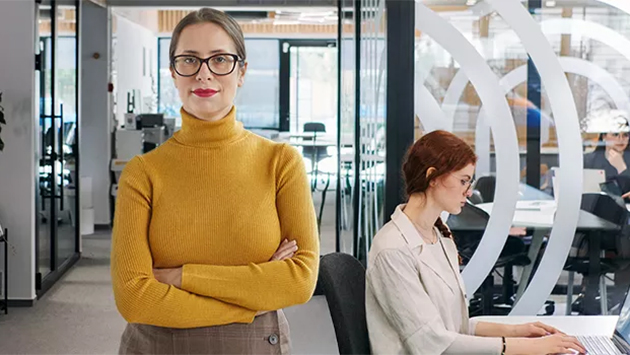 A person wearing glasses and a mustard-yellow turtleneck stands with arms crossed in the foreground of a modern open office, looking toward the camera with a calm expression. Behind them, another person works at a laptop near a glass partition etched with large concentric-circle graphics, with more colleagues at desks in the background.