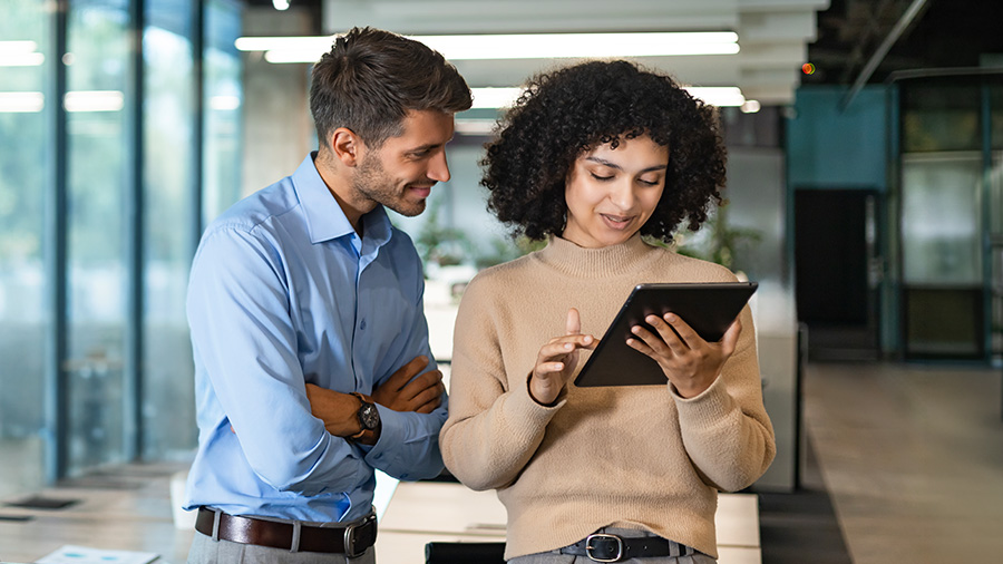Two coworkers stand together in a modern office hallway while looking at a digital tablet. The woman holds and operates the tablet, and the man stands beside her with folded arms, attentively watching the screen. Large glass windows and contemporary interior details create a bright, professional environment. Their relaxed posture and smiles suggest a friendly, collaborative work discussion.
