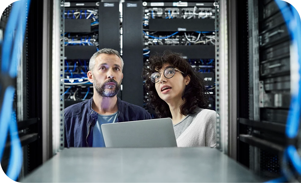 Two people stand together in a data center aisle lined with server racks, looking at a laptop one of them holds. One wears a dark sweater and the other a white top with glasses; both have focused expressions.