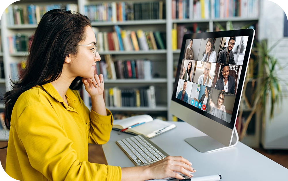 A person with long dark hair in a yellow top sits in profile at a desk, looking at a large monitor displaying a video call with a grid of participants. A keyboard rests in front of them and bookshelves fill the background.