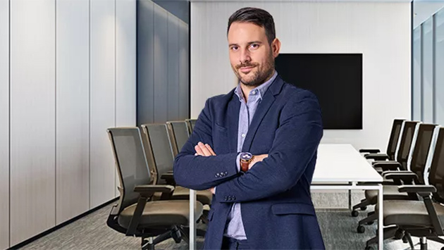 A person with a beard in a navy blazer over a blue shirt stands with arms crossed in a modern conference room, smiling toward the camera. A long white table with brown chairs and a large blank wall display fill the background.