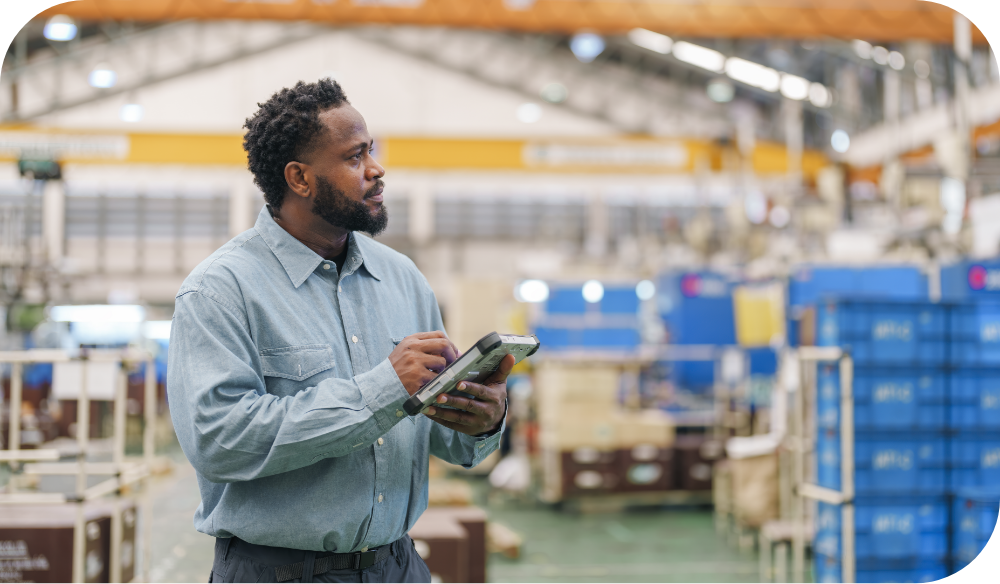A person wearing a gray button-down shirt stands in a large industrial facility, holding a tablet and looking to the side with a focused expression. Blue industrial equipment and overhead cranes are visible in the background.