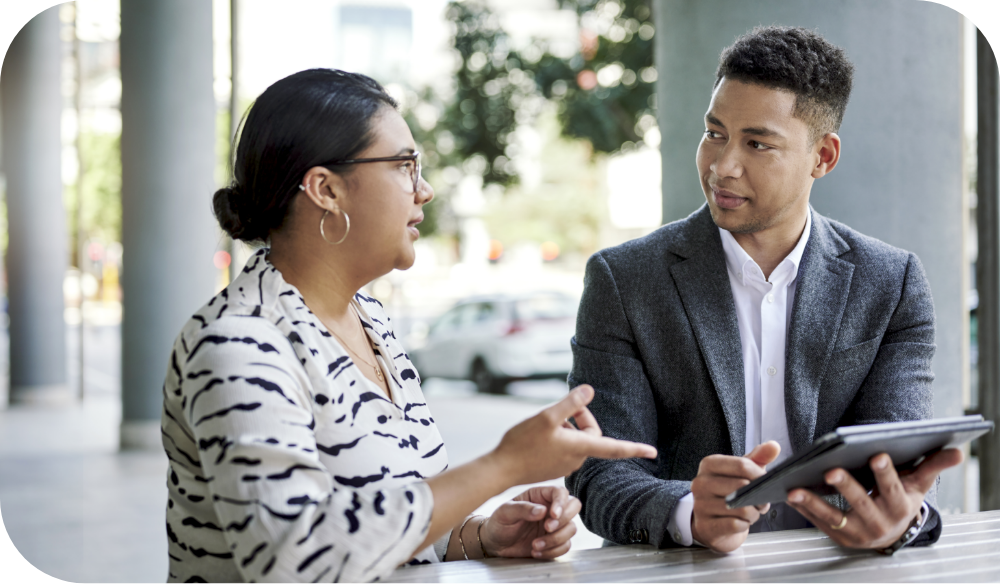 Two people sit outdoors at a table in an urban setting, engaged in conversation. One person wearing glasses and a patterned blouse gestures while speaking, and the other wearing a gray blazer holds a tablet and listens.