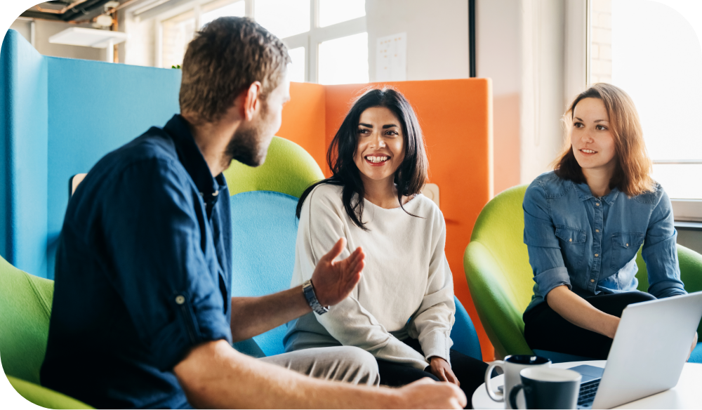 Three people sit close together in a bright, colorful office lounge having a relaxed conversation. One person in a denim shirt gestures while talking, and a second person in a light sweater smiles and gestures in response, while a third looks on. Green lounge chairs and orange and blue dividers are visible in the background.