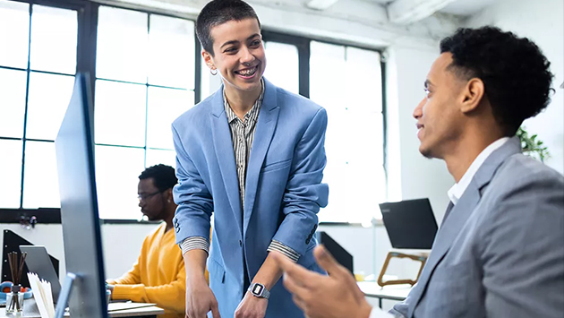 Two people stand facing each other in a modern open office, smiling and engaged in conversation. One person wearing a light blue blazer leans against a desk, and the other wearing a gray blazer holds a smartphone. A third person is visible working at a desk in the background.