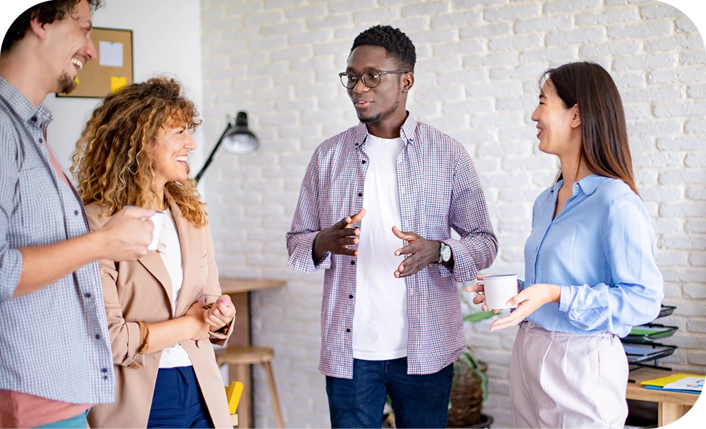 Four people stand in a group in a bright room with a white brick wall, engaged in casual conversation. They hold notebooks and cups in a creative or collaborative workspace setting.