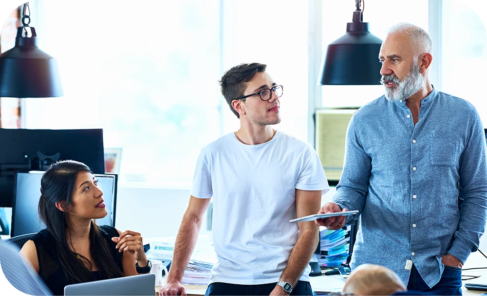 Three people are gathered around a desk in a modern office with overhead industrial lighting. One is seated at a computer monitor while the other two stand nearby, all engaged in a collaborative discussion.
