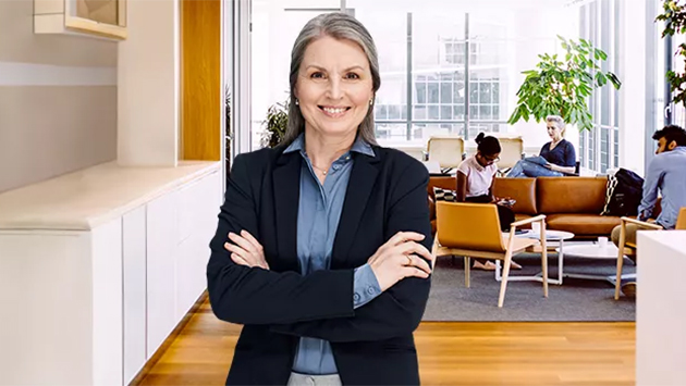 A person with shoulder-length gray hair wearing a black blazer and blue shirt stands with arms crossed and a warm smile in a modern office lobby. In the background, several people are seated in a lounge area near large windows and a potted plant.