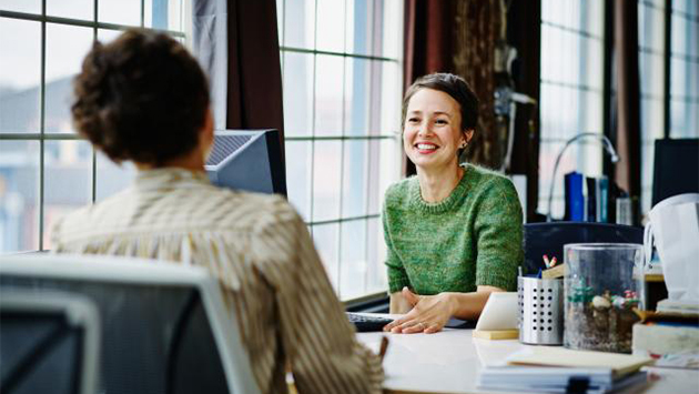 Two people stand facing each other in a bright modern office space, engaged in conversation. The person on the left holds an open laptop in one arm while gesturing with the other hand, wearing a light-colored casual button-down shirt and smiling attentively. The person on the right stands with arms relaxed, dressed in dark business-casual clothing, listening with a slight smile. Behind them, out-of-focus glass partitions, desks, and warm overhead lighting fill the background.
