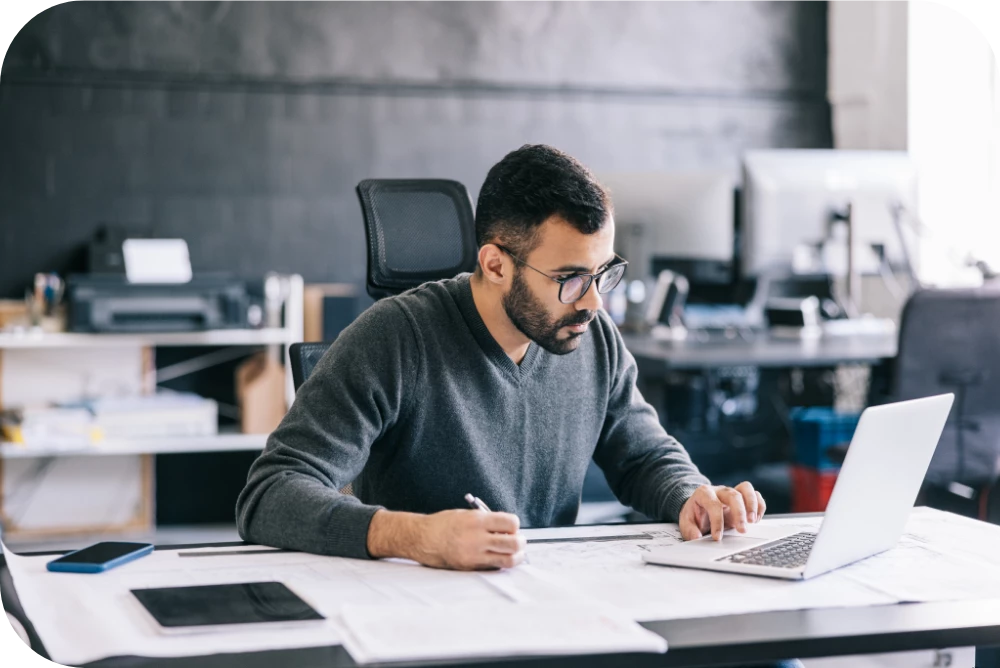 A person with a beard and glasses, wearing a gray sweater, sits at a desk writing notes beside an open laptop and printed documents in a bright office with shelves in the background.