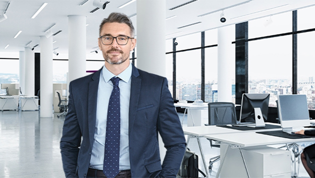 A person with gray hair, a beard, and glasses, wearing a navy suit with a light blue shirt and patterned tie, stands smiling with hands in pockets in a bright open office overlooking a city skyline through large windows.