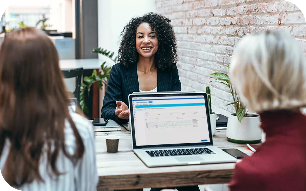 Three people sit around a conference table during a business meeting. A laptop displaying a software dashboard is in the foreground, while one participant leads the discussion. The meeting takes place in a modern office with a brick wall and indoor plants.