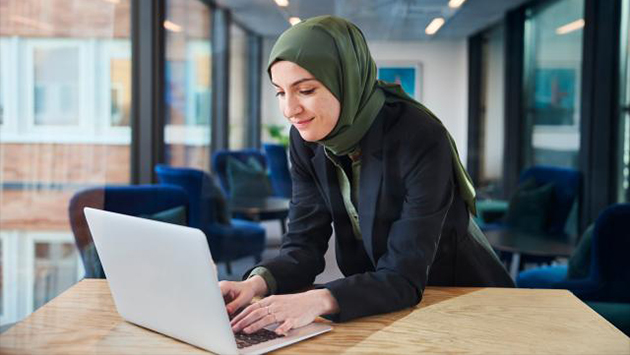 A person wearing a green headscarf and a dark blazer leans forward over a wooden table, typing on a laptop with a slight smile. Blue lounge chairs and large windows with a building exterior fill the bright background.