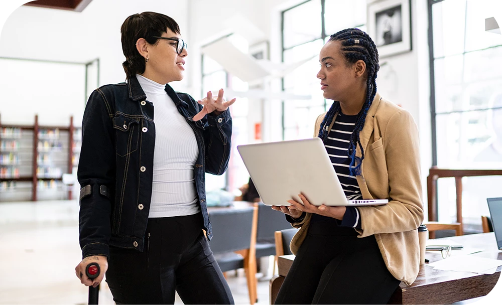 Two people stand talking in a bright library or open office. One wears a denim jacket and gestures while holding a sheet of paper; the other wears a striped top and blazer and holds an open laptop. Bookshelves and large windows fill the background.
