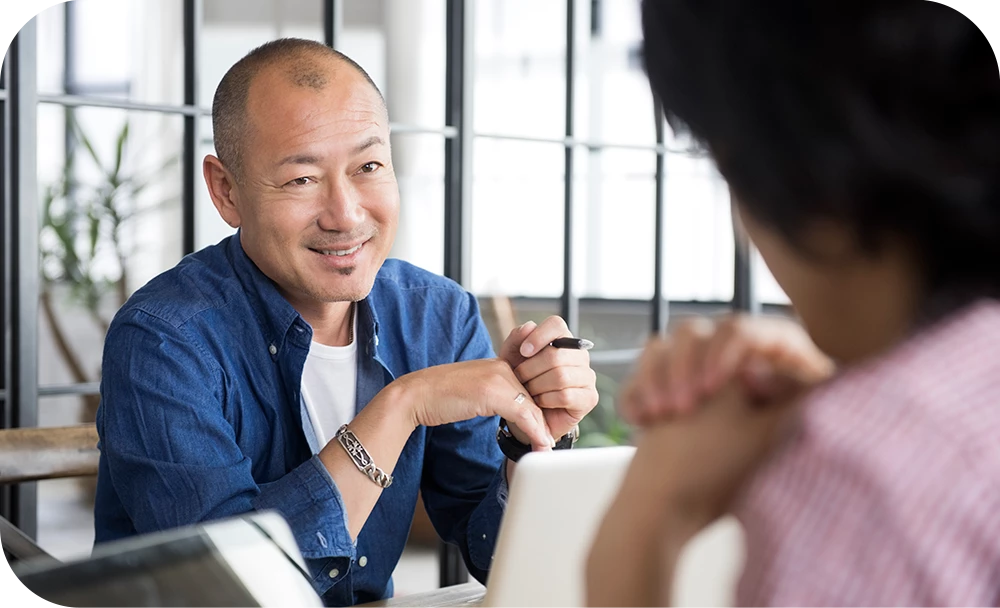 A person with short hair, wearing a blue button-up shirt over a white tee, sits at a table smiling and resting their clasped hands on the surface while talking with another person seen from behind in the foreground. The setting is a bright, casual space with soft greenery in the background.