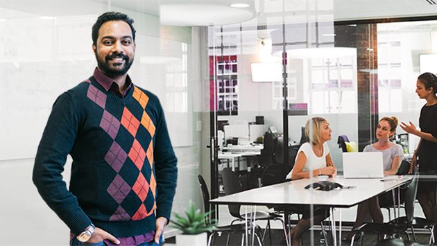A person wearing a dark argyle sweater in shades of purple and orange over a checked shirt stands smiling in the foreground of a modern office, one hand resting at the waist. Behind a glass partition, three colleagues gather around a table with a laptop in a meeting.