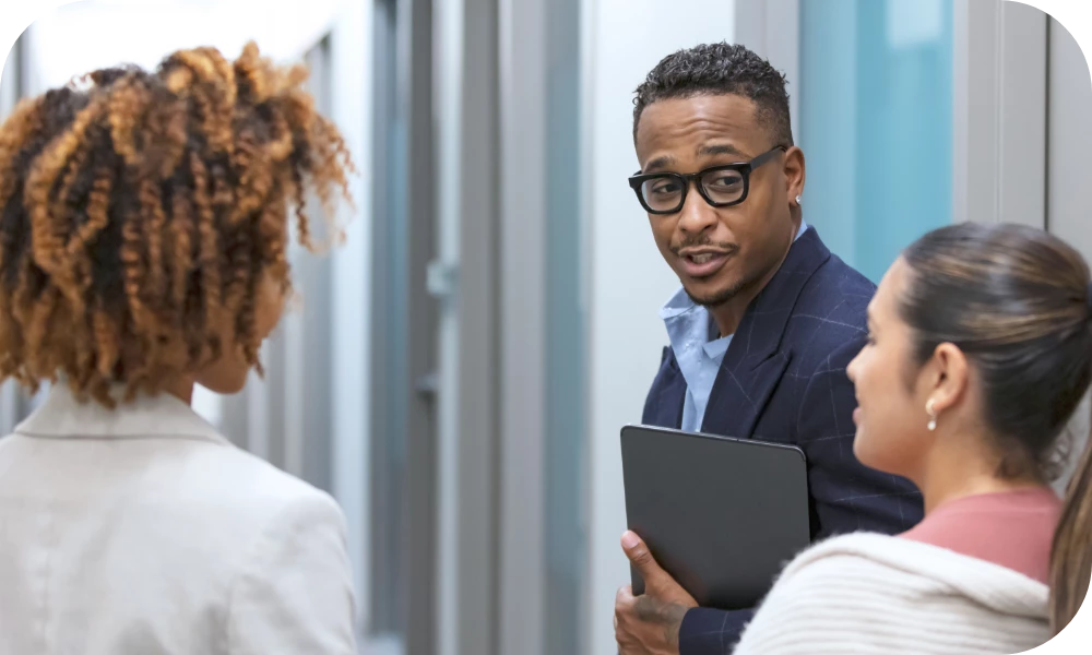 Three people stand in a hallway in conversation. A person in the center, wearing glasses and a dark blazer, holds a tablet and speaks while two others listen, one on each side. The setting is a bright modern office corridor.