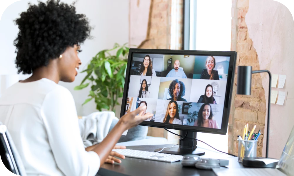 A person with curly hair, wearing a white top, sits at a desk facing a monitor that displays a video call grid of several participants. The person gestures toward the screen in a bright home office with a plant and a pen holder on the desk.