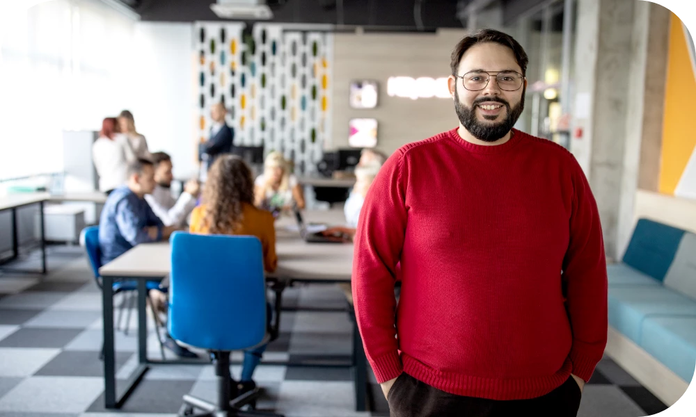A person with short dark hair, a beard, and glasses, wearing a red sweater, stands in a modern coworking space with hands clasped and smiles toward the camera. Behind them, several people sit at tables and a patterned wall and blue accent chairs are visible.