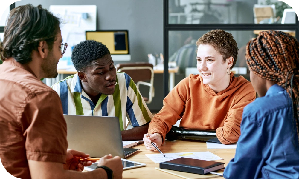 Four people sit around a table in a bright office collaborating. One person in an orange shirt and another in a yellow sweater look at a laptop and tablet on the table, while the others participate in the discussion.