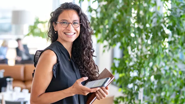 A person with long curly hair and glasses, wearing a sleeveless black top, stands in a bright office holding a smartphone and a notebook, smiling at the camera. Lush green foliage and a lounge area are visible in the background.