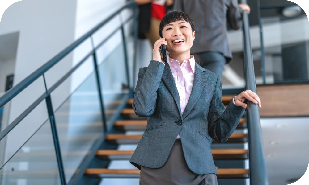 A person in a gray suit stands on an interior staircase smiling while talking on a smartphone, holding the railing with one hand. Another person is partially visible ascending the stairs in the background.