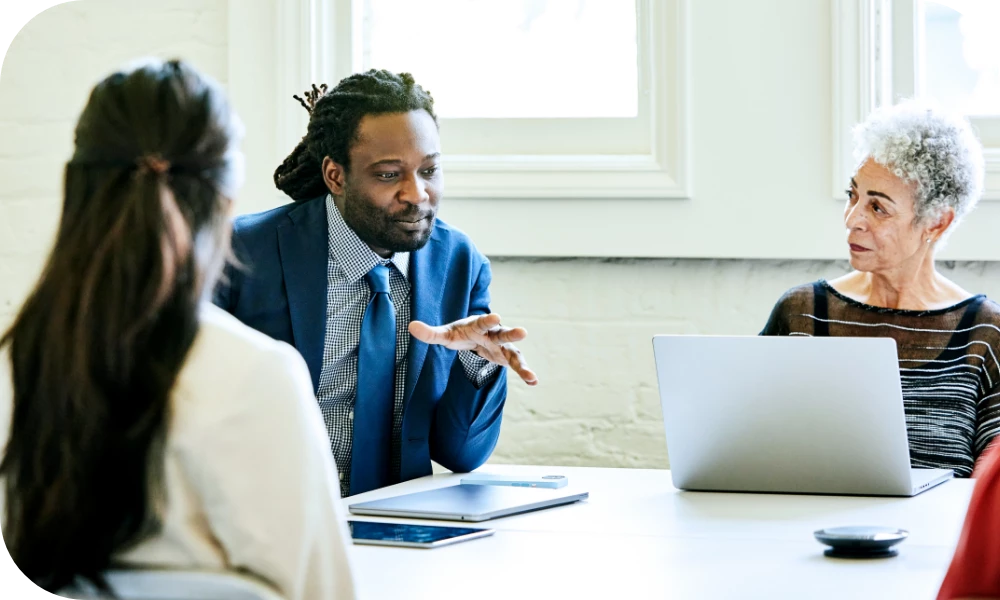 Three people sit at a table in a meeting. A person in a dark suit and blue tie gestures while speaking, a person with gray hair listens beside an open laptop, and a third person is seen from behind in the foreground. The setting is a bright, modern office.