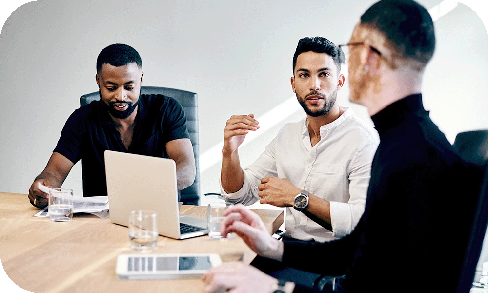 Three people sit around a wooden conference table in a meeting. The person in the center, wearing a white shirt, gestures with one hand while speaking. Laptops, a tablet, water glasses, and a bottle rest on the table. Bright daylight comes from a window behind them.