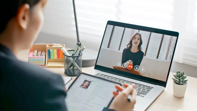 Seen from over the shoulder of a person with dark hair wearing a dark blazer, a laptop on a desk shows a video call with another person who is speaking. The foreground person holds a stylus and takes notes on a tablet. The desk also holds a mesh cup of pens, a stack of sticky notes, a desk lamp, and a small potted succulent, with a bright window in the background.