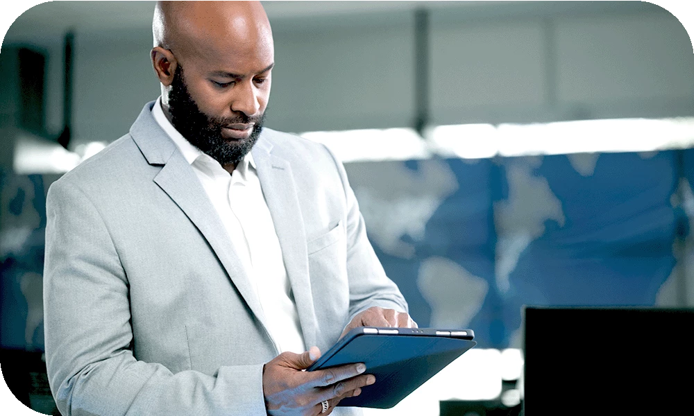 A person with a beard in a light gray suit stands in a bright office, looking down at a tablet held in both hands with a focused expression. A large blurred world map display glows in the background.