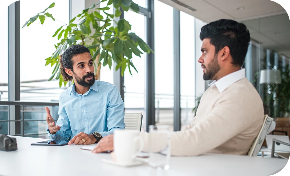 Two people sit across from each other at a table in a bright, modern office with large windows and plants. One gestures expressively while speaking; the other listens attentively. Coffee cups rest on the table between them.