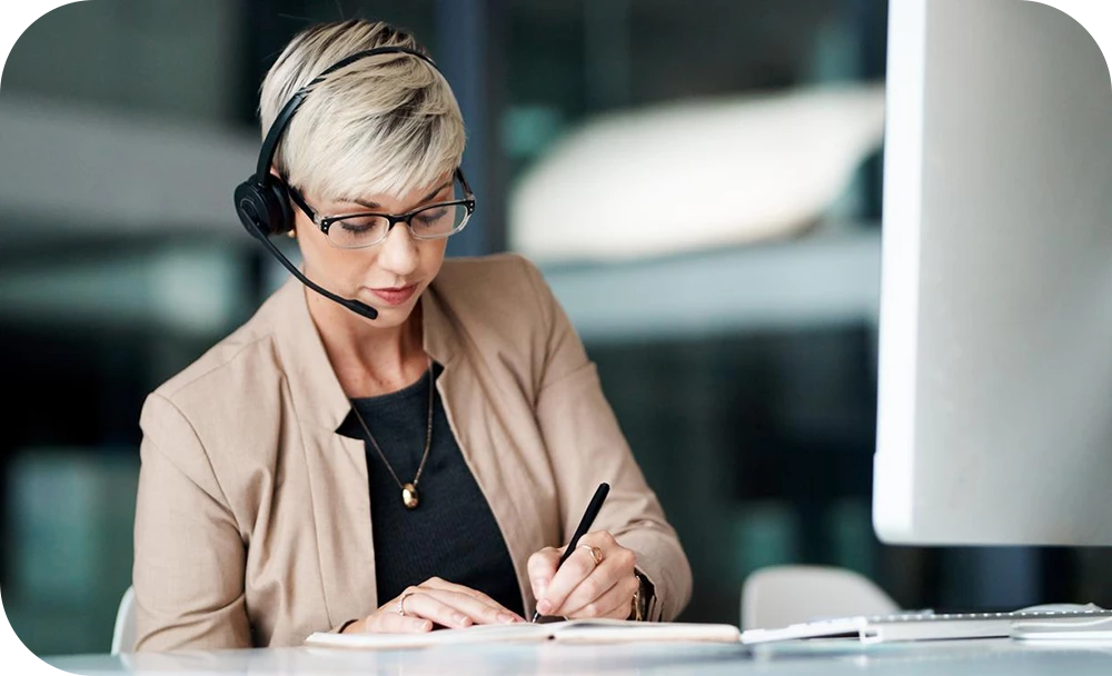 A person wearing a headset sits at a desk in front of a monitor, writing notes in a notebook with a focused expression. They wear a beige blazer and a dark necklace in a professional office setting.