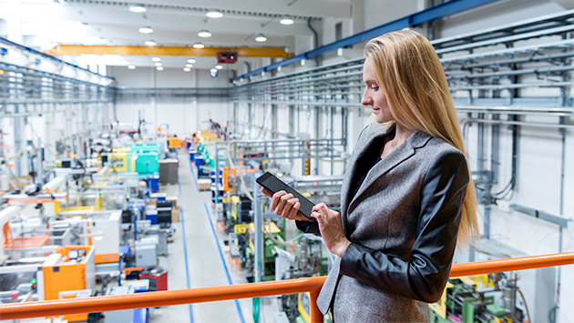 A person with long hair, wearing a gray business blazer, stands on an elevated walkway behind an orange safety railing inside a large industrial factory. The person holds a tablet in both hands, looking down at it with a slight smile. Below and behind them, the factory floor stretches into the distance with rows of colorful manufacturing machinery, pipes, ductwork, and overhead lighting under a high ceiling.