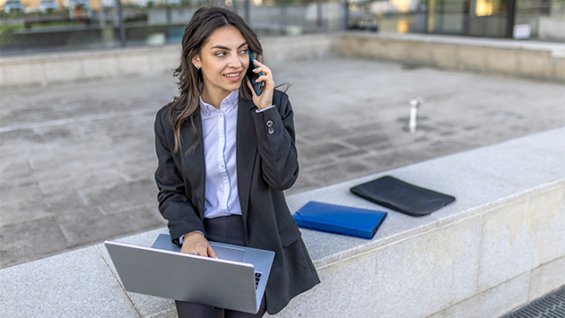 A person with long dark hair, wearing a dark blazer over a light blue collared shirt, sits on a concrete ledge outside a modern building. They balance an open laptop on their lap with one hand while holding a smartphone to their ear with the other, looking off to the side with a slight smile. A blue folder and a dark tablet sleeve rest on the ledge beside them.