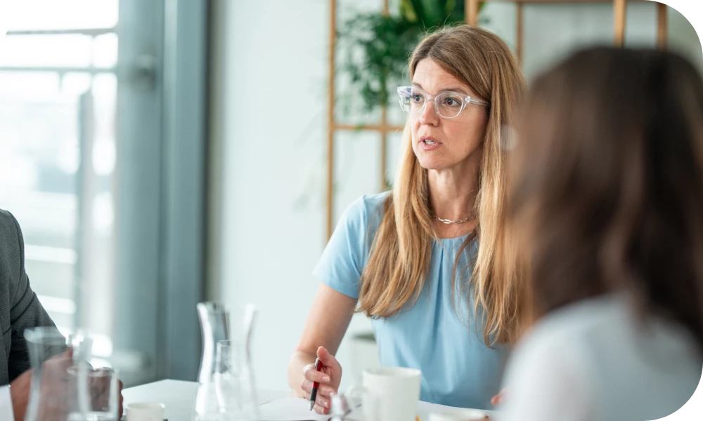 A person with long hair, wearing a light blue top and glasses, sits at a table in a bright office listening attentively during a meeting. Cups and a water carafe are on the table, and another person is visible from behind in the foreground.