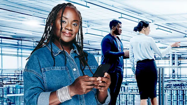 A person with long braided hair, wearing a denim jacket, stands in the foreground of a data center holding a smartphone and smiling at the camera. Behind them, two other people stand near server racks, one gesturing toward the equipment.