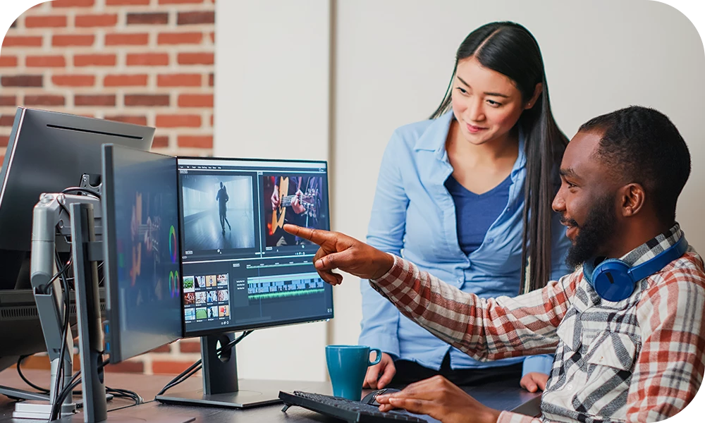 Two people seated side by side at a dual-monitor workstation in a creative studio. One person points at a screen displaying video editing software with footage visible while both are focused on the monitors. A coffee cup and desk accessories are visible.