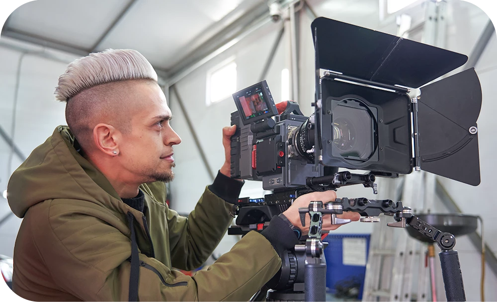 A person wearing a jacket operates a professional cinema camera mounted on a rig outdoors, looking through the viewfinder in an active production environment.
