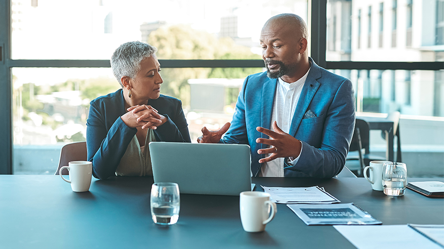 Two business professionals in an office setting seated in conversation while both looking at one laptop. 