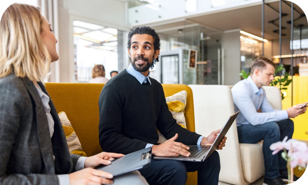 Two people sit facing each other in a bright office lounge with yellow seating, each with a laptop. One person, wearing a dark blazer over a black top, gestures while talking and smiles slightly. The other person, seen from behind with light hair, holds a laptop. In the background another person works on a laptop near large windows.