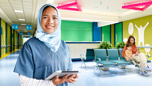 A person wearing a light blue head covering and blue medical scrubs over a white long-sleeve top stands in the foreground holding a tablet and smiles slightly toward the camera. Behind them is a brightly lit clinic waiting area with green cabinetry, blue flooring, teal waiting chairs, and pink geometric ceiling fixtures. In the background another person sits on a bench with a young child, and a white wall graphic of a figure with raised arms is visible.