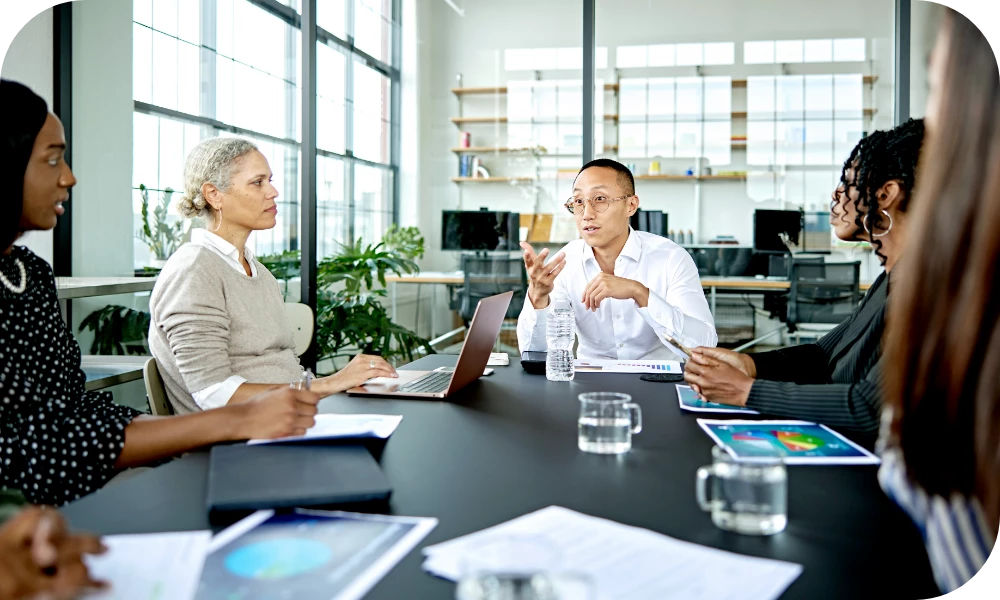 Several people sit around a conference table in a bright, modern meeting room. A person in the center gestures while speaking, and the others listen. Laptops, papers with charts, and water glasses are on the table.