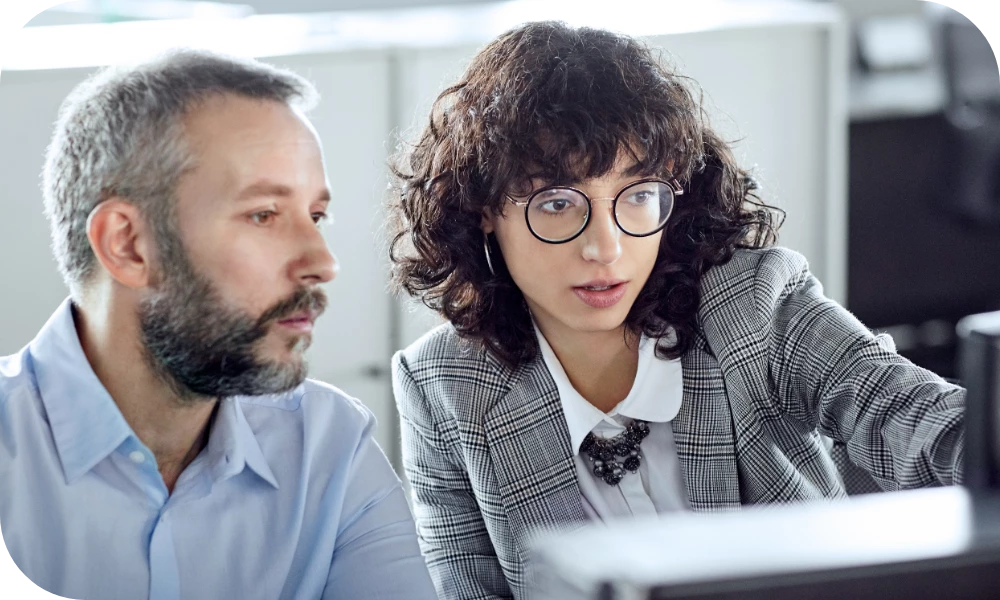 Two people sit side by side looking at a computer screen in an office. One has a beard and wears a dark top; the other has curly hair and glasses and wears a checked blazer. Both look focused on the screen in front of them.