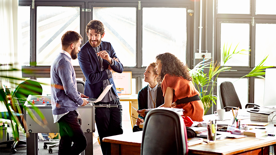 A group of colleagues gathered in a bright office, discussing work around a laptop and desk. 