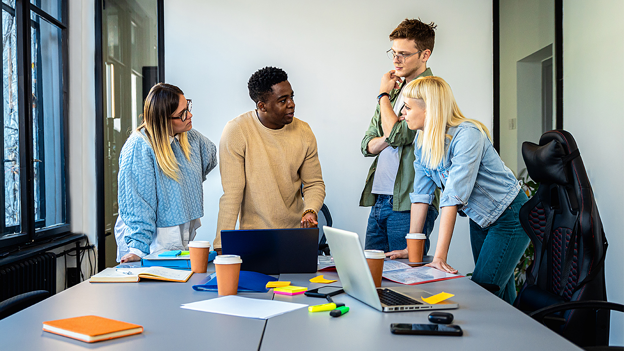 Team members gathered around a conference table, collaborating over laptops, notes, and documents in a modern office.
