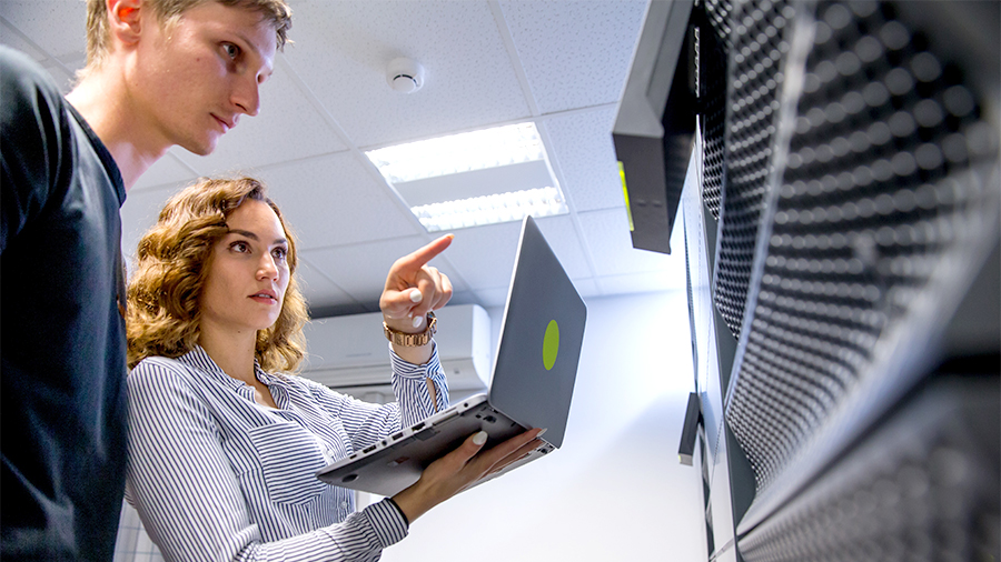 Two IT professionals monitoring server infrastructure, with one pointing at equipment while holding a laptop. 